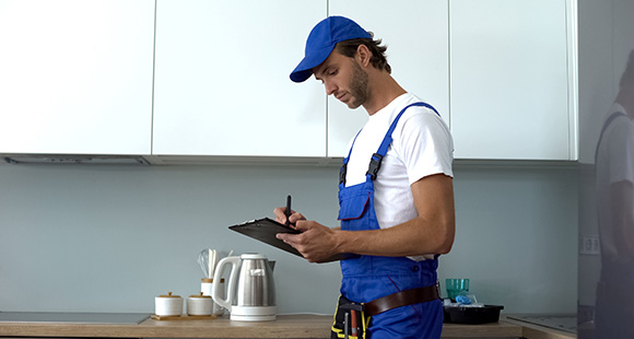 a man wearing a blue hat and overalls writing a report on electrical safety in a kitchen