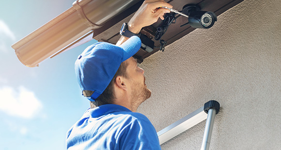 a man with a blue hat and uniform on a ladder installing a security alarm on a house